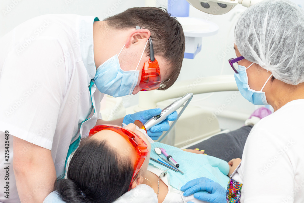 girl patient at the reception at the dentist. treatment of carious tooth. the girl lies on the dental chair with his mouth open. A dentist and his assistant are treating a tooth girl patient at the reception at the dentist. treatment of carious tooth. the girl lies on the dental chair with his mouth open. A dentist and his assistant are treating a tooth