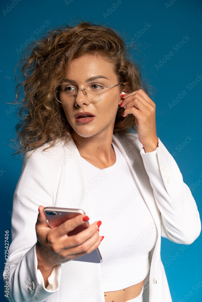successful happy curly woman in white shirt. With blue background. Beautiful business woman, with earphones and smartphone in hands