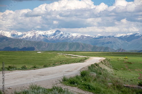 Country road among green fields and hills against the backdrop of mountains with snow-capped peaks