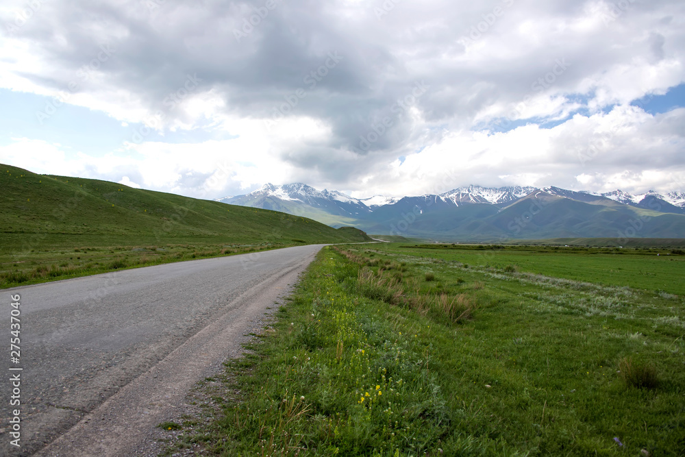 Country road among green fields and hills against the backdrop of mountains with snow-capped peaks