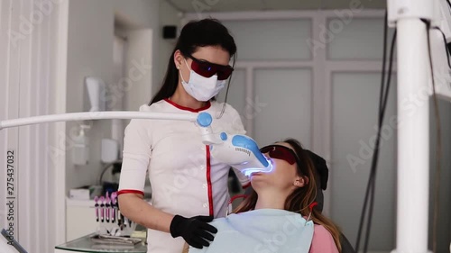 Procedure of teeth whitening in dentistry clinic. Young woman in protective glasses sitting on dentist chair. Doctor holding modern device, ultraviolet lamp with blue light. Female dentist in white photo