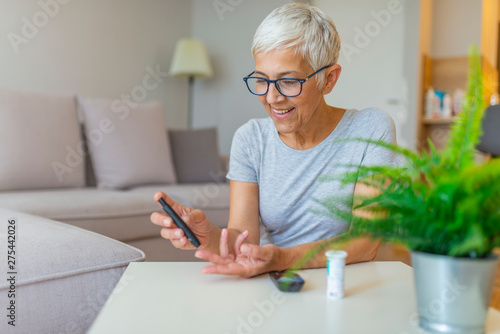 Happy mature woman with glucometer checking blood sugar level at home. Woman testing for high blood sugar. Woman holding device for measuring blood sugar