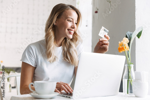 Cheerful young pretty blonde woman student sitting in cafe using laptop computer drinking coffee holding credit card.