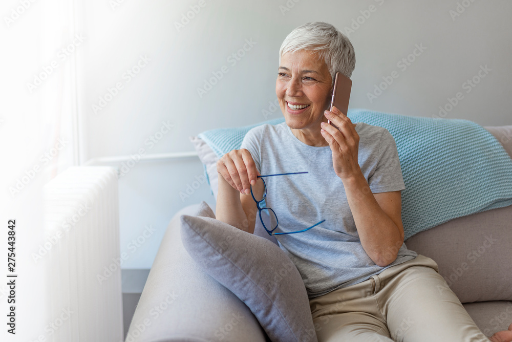 Senior woman talking on her mobile phone. Senior woman has a happy conversation at cellphone. Smiling senior woman using phone sitting on couch at home. Senior woman talking on her mobile phone. Senior woman has a happy conversation at cellphone. Smiling senior woman using phone sitting on couch at home.