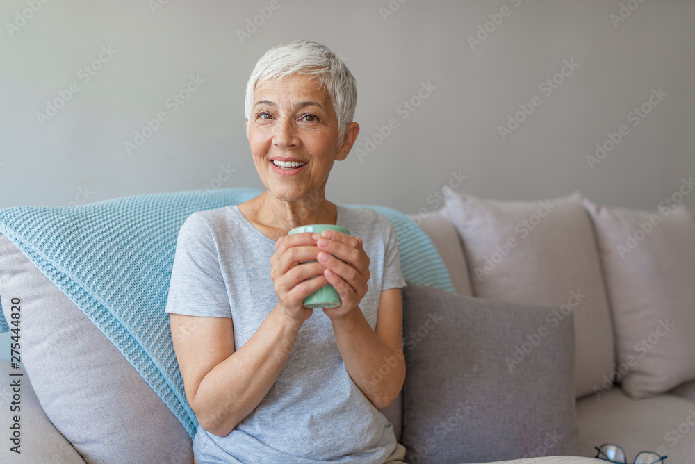 Happy senior woman sitting on a couch while she is drinking a cup of coffee. Portrait of smiling senior woman drinking coffee at home while looking at camera. Happy senior woman sitting on a couch while she is drinking a cup of coffee. Portrait of smiling senior woman drinking coffee at home while looking at camera.