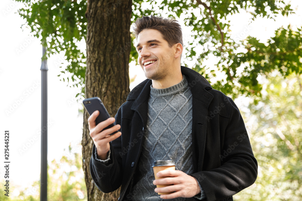 Handsome happy man in casual clothing walking outdoors in green park using mobile phone drinking coffee. Handsome happy man in casual clothing walking outdoors in green park using mobile phone drinking coffee.