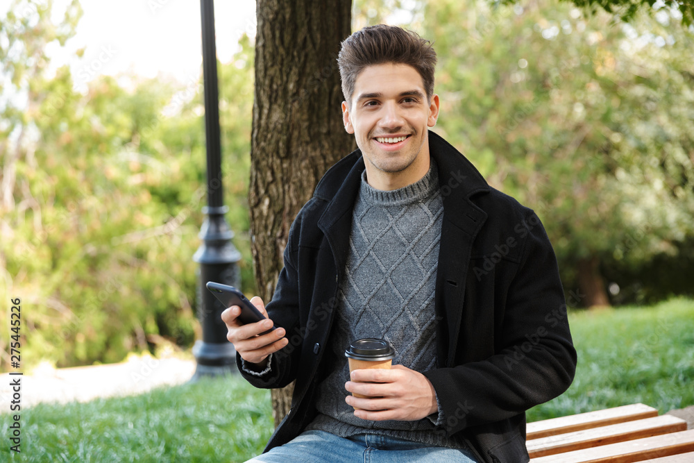Handsome cheerful young happy man in casual clothing walking outdoors in green park using mobile phone drinking coffee. Handsome cheerful young happy man in casual clothing walking outdoors in green park using mobile phone drinking coffee.