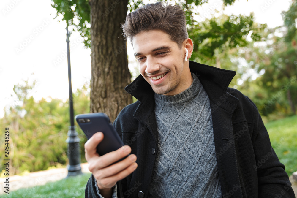 Young cheerful man in casual clothing walking outdoors in green park using mobile phone listening music with earphones. Young cheerful man in casual clothing walking outdoors in green park using mobile phone listening music with earphones.