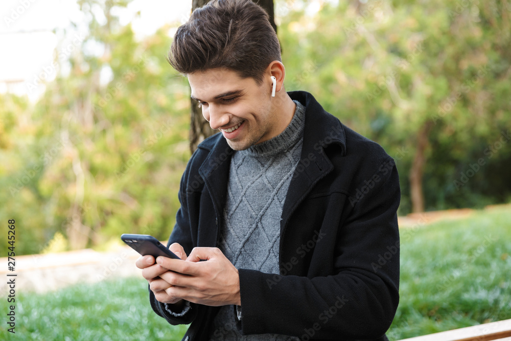 Smiling handsome young cheerful man in casual clothing walking outdoors in green park using mobile phone listening music with earphones. Smiling handsome young cheerful man in casual clothing walking outdoors in green park using mobile phone listening music with earphones.