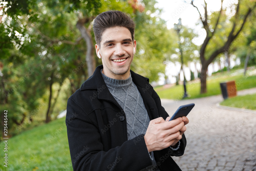 Smiling handsome young cheerful man in casual clothing walking outdoors in green park using mobile phone listening music with earphones. Smiling handsome young cheerful man in casual clothing walking outdoors in green park using mobile phone listening music with earphones.
