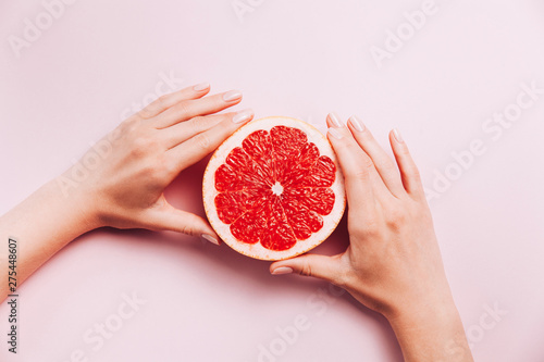 Female's hands holding grapefruit on pink background with tropic plants and leaves.