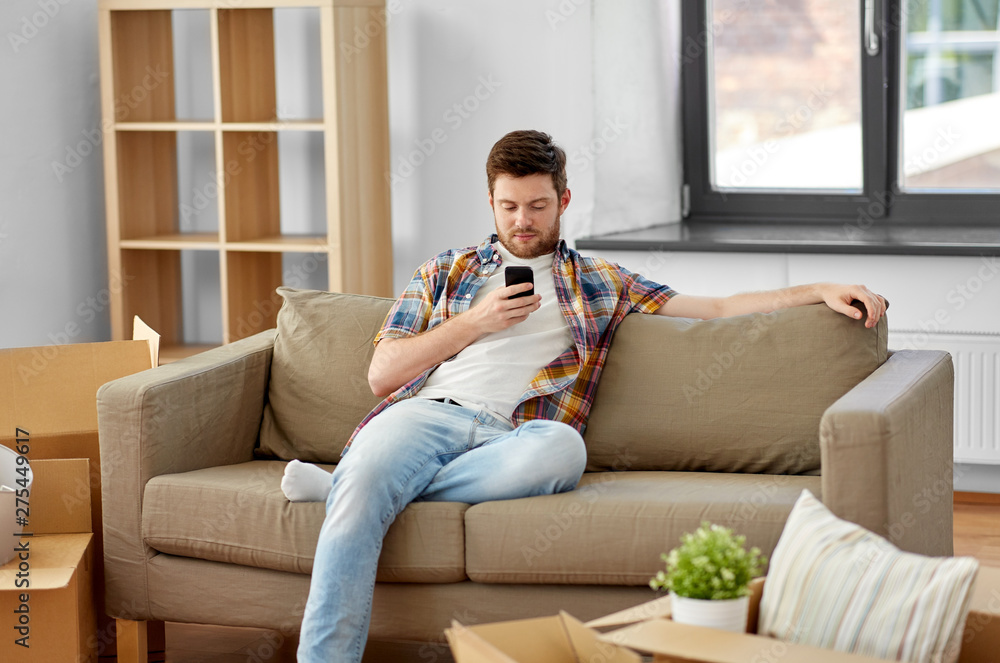 moving, people and real estate concept - happy man with smartphone and boxes sitting on sofa at new home