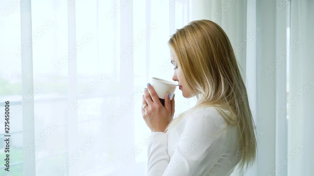 side view of young blonde woman looking through window and drinking coffee side view of young blonde woman looking through window and drinking coffee