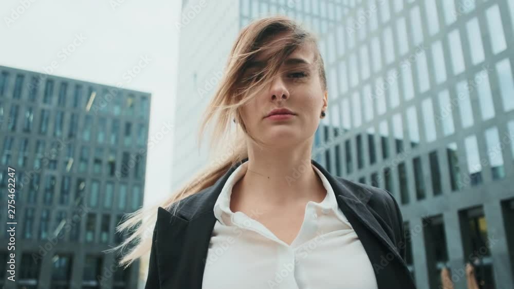 Portrait of young business woman in suit with blonde hair blowing in wind walking on the urban business district street. Skyscraper background. Business, finance, banking. Girl looking at camera. Portrait of young business woman in suit with blonde hair blowing in wind walking on the urban business district street. Skyscraper background. Business, finance, banking. Girl looking at camera.