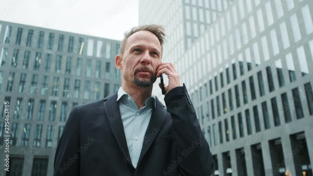 Successful business man talking on smartphone while walking on the urban business district street. Skyscraper background. Business, finance, banking. Successful business man talking on smartphone while walking on the urban business district street. Skyscraper background. Business, finance, banking.