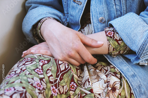 mid section of young woman wearing denim jacket over summer dress with floral pattern - close-up of female hands folded in her lap