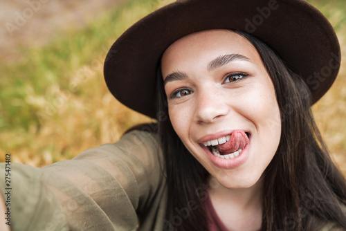 Photo closeup of cheerful woman wearing hat showing her tongue at camera while taking selfie photo outdoors