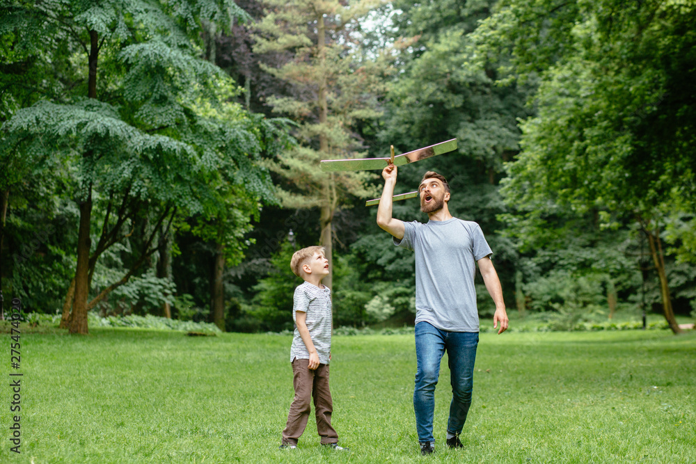 Fototapeta premium Dad and son together launch model airplane outdoors, both man and boy are looking cheerfully at plane. Father and son spending good time together, family relationships concept. Full leight