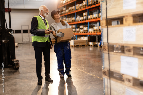 Mature businessman and warehouse employee communicating while walking through industrial building.