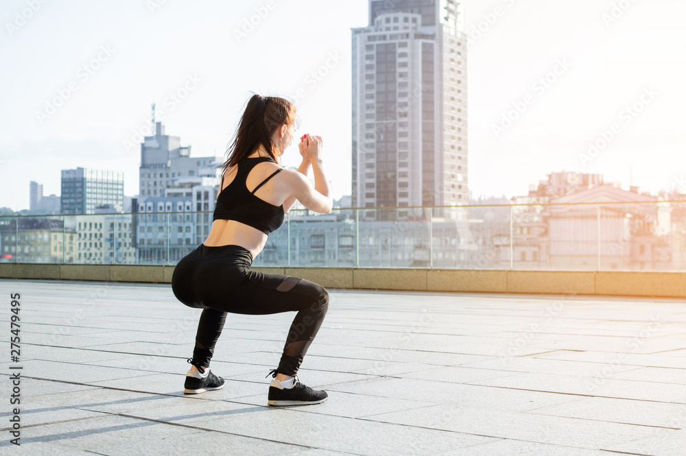 athletic girl crouches on the background of the city, she does an exercise on the street, fitness woman train in the morning