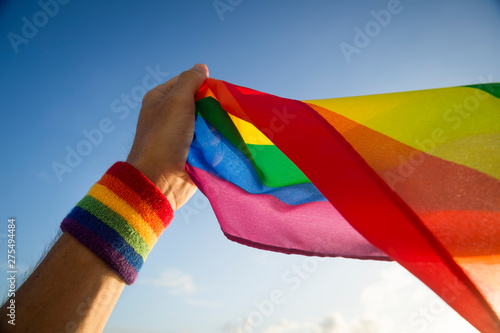 A hand with rainbow color wristband waving gay pride flag backlit in the wind against golden sunset sky