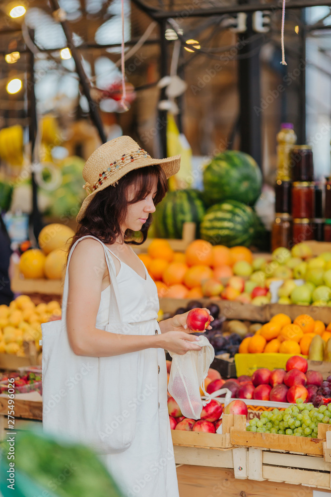 Obraz premium Young Woman puts fruits and vegetables in cotton produce bag at food market. Reusable eco bag for shopping. Zero waste concept.
