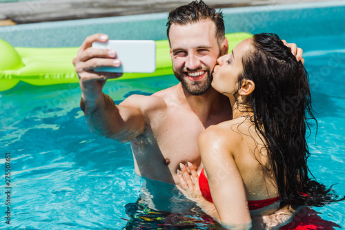 selective focus of woman kissing cheek of happy man taking selfie in swimming pool