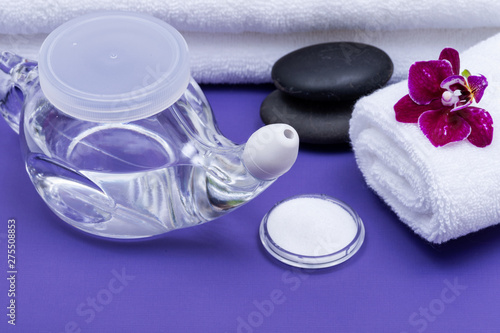 Spa purple background with Neti Pot, pile of Saline, rolled up White Towels, stacked Basalt Stones and Orchid Flower. Sinus wash. Nasal irrigation.
