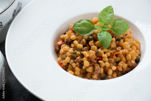 Close-up of fregola pasta with bell pepper, capers and fresh green basil served in a white plate