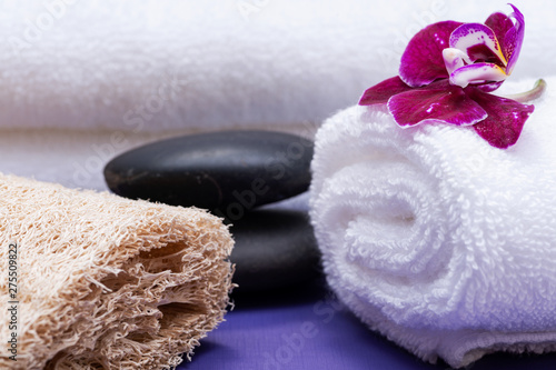 Spa Wellness Concept. Natural Loofah Sponge, rolled up White Towels, stacked Basalt Stones and Orchid Flower on purple background.