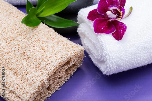 Spa Wellness Concept. Natural Loofah Sponge, rolled up White Towels, stacked Basalt Stones, Bamboo and Orchid Flower on purple background.