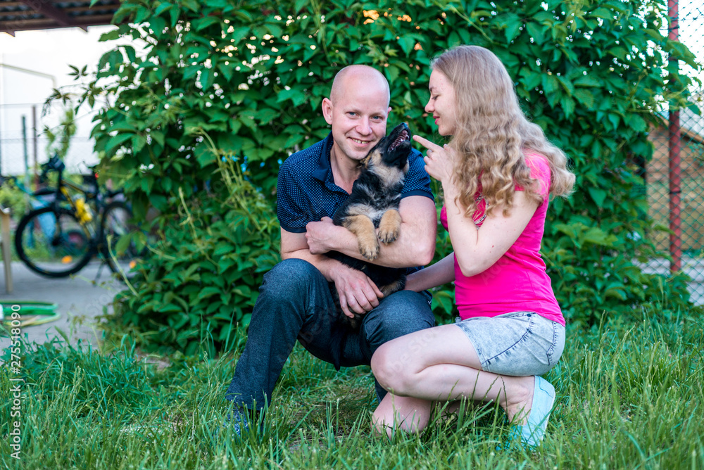 Fototapeta premium Young happy couple with a funny puppy of a German shepherd. A girl and a guy on a green lawn near the house with a new dog. Buying a pet. The joy of first meeting. Copy spase