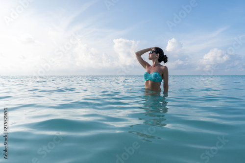 A young girl is standing to the waist in the turquoise water and looking toward the sun.