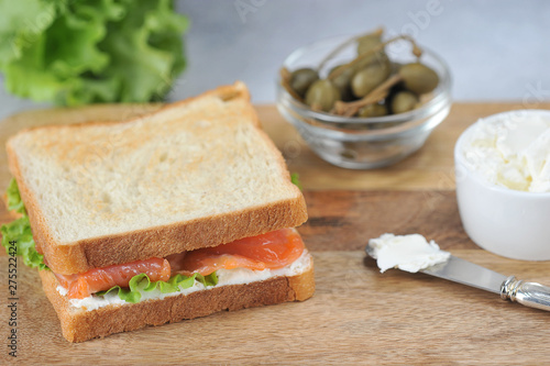 Salmon sandwich on a light wooden board. Cups with capers and soft cheese. Close-up.