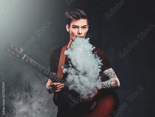 Young attractive man is playing acoustic guitar while smoking vape, making big haze.