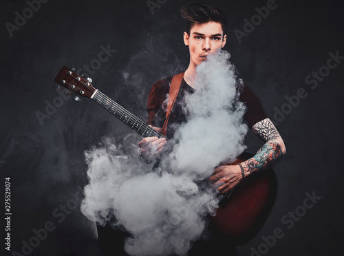Cheeky young man is smoking, making nice vapour while holdind acoustic guitar at studio.