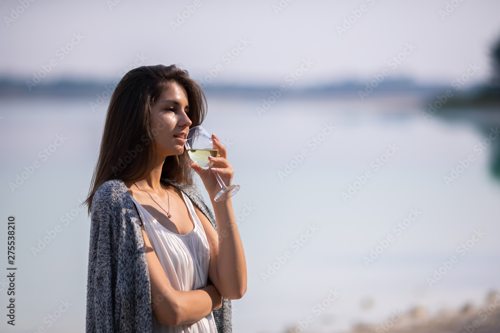 Young beautiful girl in white dress drinks wine from a glass. She is on the shore of the lake and enjoy a picnic and outdoor recreation.