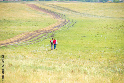 Two young girls with backpacks and photo camera make selfie on phone near rural road. Horse farm pasture with mare and foal. Small village with old houses. Summer landscape with green hills © GMars