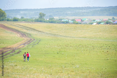 Two young girls with backpacks and photo camera make selfie on phone near rural road. Horse farm pasture with mare and foal. Small village with old houses. Summer landscape with green hills