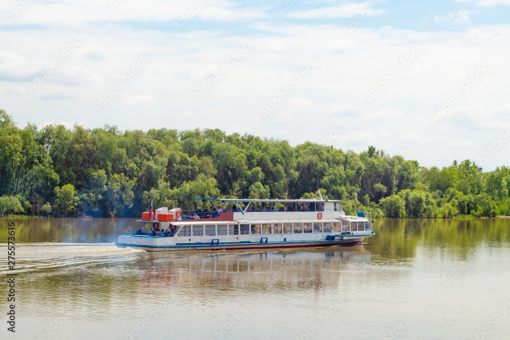 A white tourist ship is slowly sailing along the banks of the Irtysh River in Omsk in the summer against the green trees on the shore, side view - travel, tourism, rest on the water