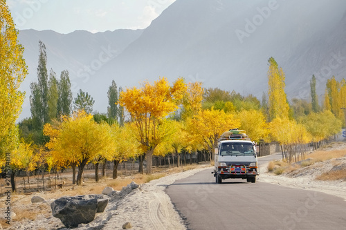 A local bus running on paved road with luggage bags on the roof with morning sunlight. Landscape view of colorful trees in autumn against mountain range in Skardu. Gilgit Baltistan, Pakistan.