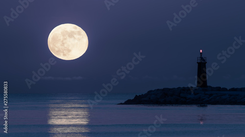 Romantic moonrise over the Mediterranean at the eastern pier in the Spanish port city of Denia. The red lighthouse flashes and the moonlight reflects in the water.