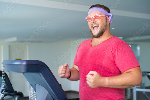 Playful fat man in a pink T-shirt and pink glasses is engaged in fitness in the gym.