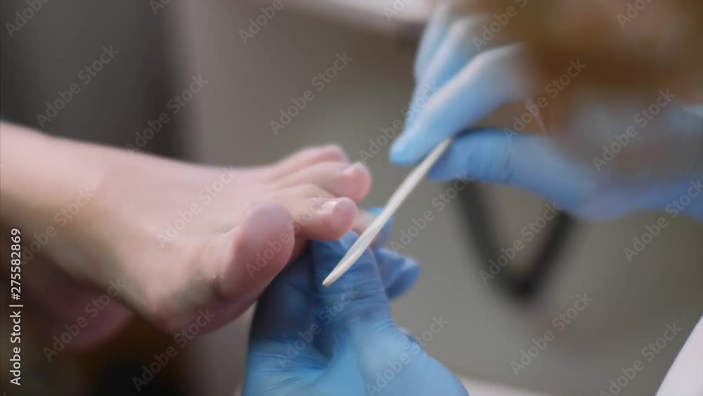 Pedicure in beauty salon. Pedicurist master in blue gloves is modeling nails on client's toes using nail file. She's dusting off toe with a brush, foot closeup.