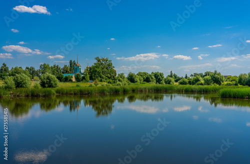 Blue sky with rare clouds reflected from the surface of the pond on the shore of which stands a blue Church surrounded by greenery