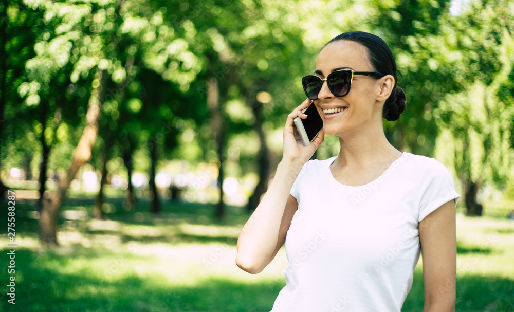 Cute smiling young woman in sunglasses is using her smart phone outdoors in city park