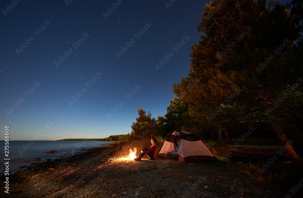 Night camping at sea shore. Attractive tourist girl sitting relaxed in front of tent at campfire under bright starry sky, enjoying beautiful view of clear blue water. Tourism, active lifestyle concept - obrazy, fototapety, plakaty 