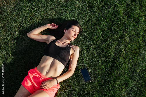 Portrait of a happy young fitness brunette woman laughing and resting on grass and listening to music on your smartphone after workout and smiling. Sport in open air and health care concept.