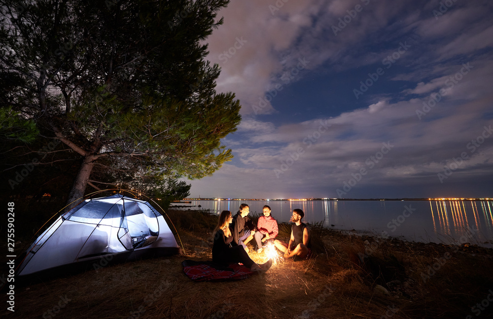 Φωτογραφία Group of four young tourists, men and women having a rest on sea shore at campfire near tourist tent