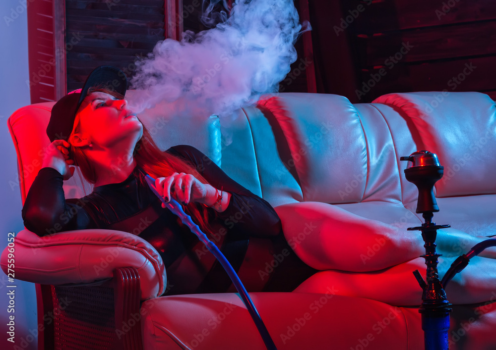 sexy redhead woman in black bodysuit sitting on a sofa and smokes hookah, girl blows out big smoke, red blue tones